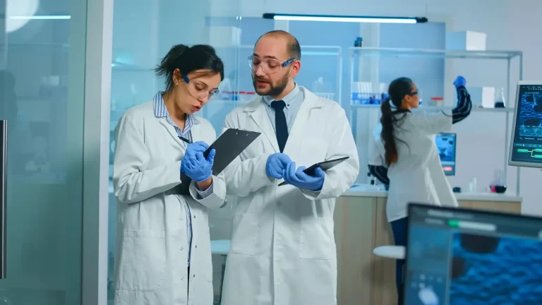 Two scientists in lab coats and safety goggles discuss notes on clipboards in a laboratory. A third scientist works with equipment in the background, and computer screens display data. The atmosphere is focused and professional.