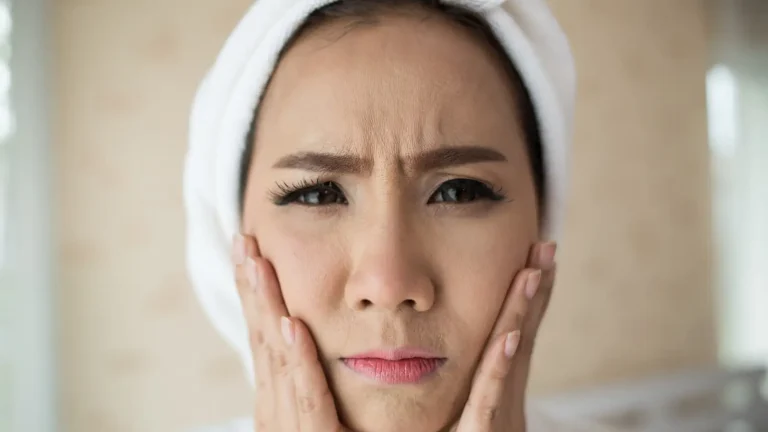 A person with a towel wrapped around their head looks directly at the camera, pressing their hands against their cheeks. They have a concerned expression, with furrowed brows, and appear to be indoors.