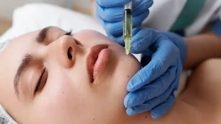 A person is lying down with eyes closed while someone wearing blue gloves administers an injection to their chin. The person receiving the injection is wearing a hair cap. The setting appears clinical, suggesting a cosmetic procedure.