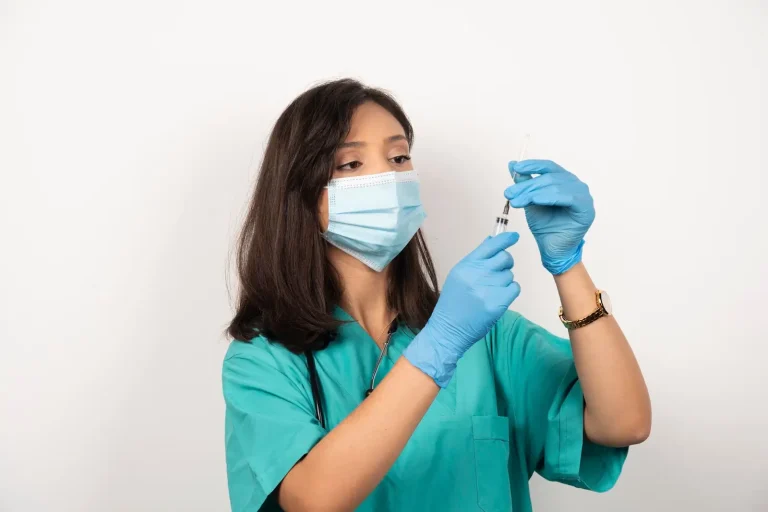 A female healthcare worker in a turquoise uniform and blue gloves, wearing a face mask, holds up a syringe and examines its contents against a plain white background. She appears focused and attentive.