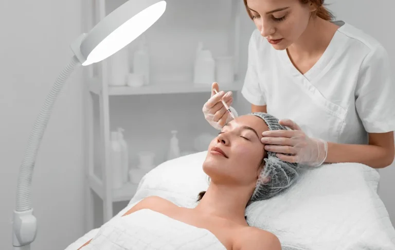A woman lies on a treatment bed with eyes closed, covered in a white towel and a hair cap. A professional, wearing gloves and a white uniform, holds a syringe, applying a cosmetic treatment. A bright lamp and shelves with products are in the background.