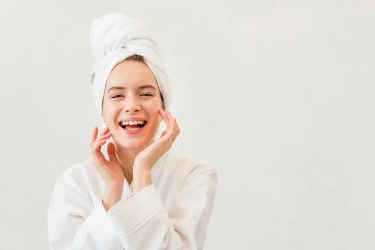 A person smiling while wearing a white bathrobe and a towel wrapped around their head, standing against a plain light background. They are touching their face with their hands, appearing relaxed and content.