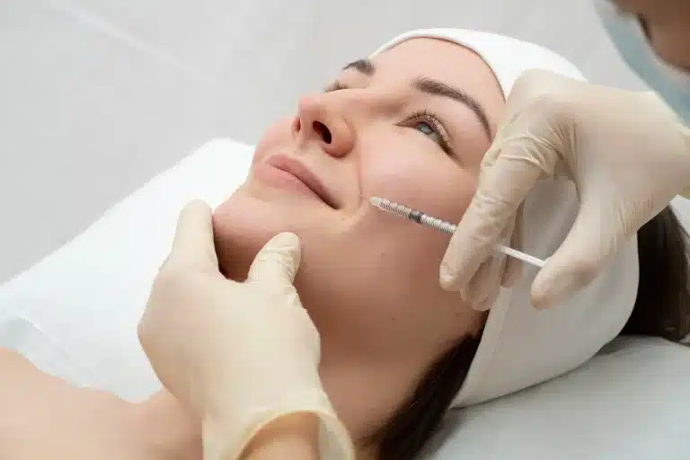 A person is lying down with a headband on, smiling slightly as a healthcare professional in gloves administers an injection to their cheek. The setting appears to be a clinical or spa environment.