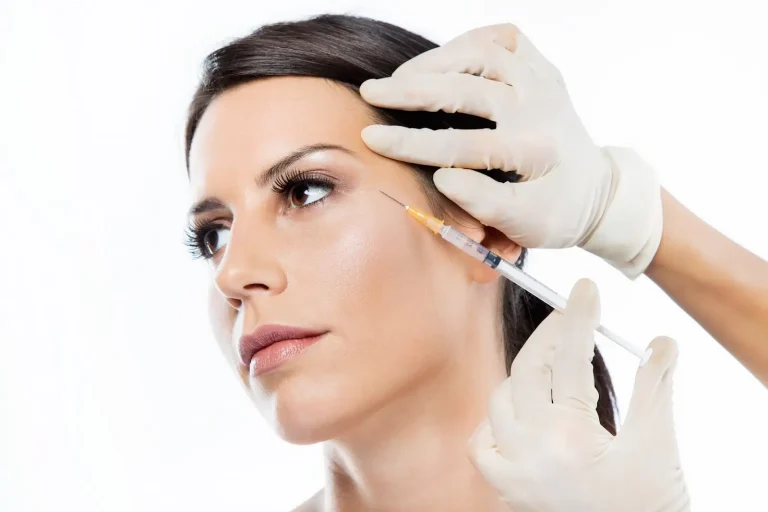 A person receives a cosmetic injection in the face. They have dark hair pulled back, and a serious expression. The practitioner's hands wear white gloves, gently holding the person's face while administering the injection. The background is white.