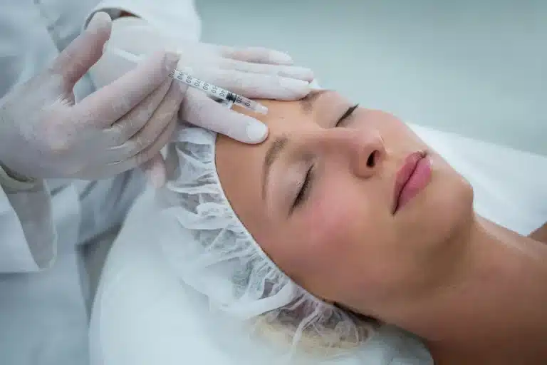 A person receiving a cosmetic injection in the forehead while lying down with closed eyes. They are wearing a hair cover and a white gown. A gloved hand holds the syringe.