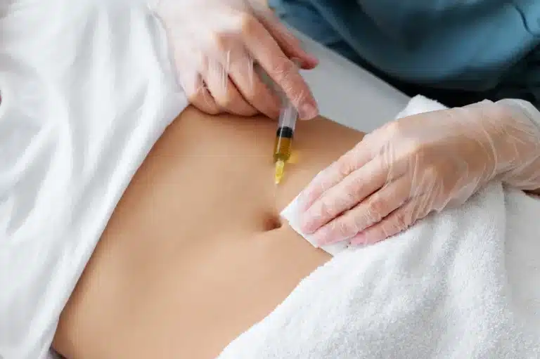 A person receives an injection in the abdominal area from a medical professional wearing gloves; a white towel covers the lower body.