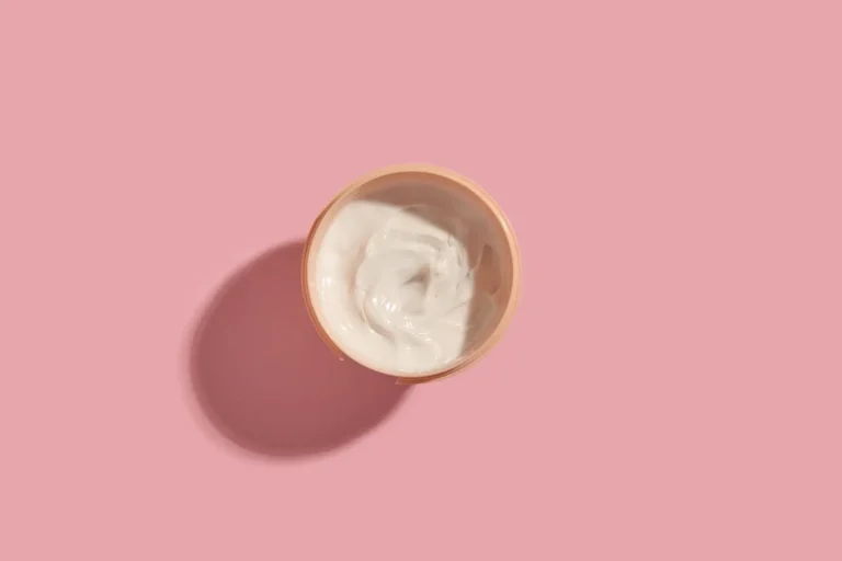 A small, open jar filled with white cream is centered on a pink background. The jar casts a soft shadow to the left, creating a simple and minimalist composition.