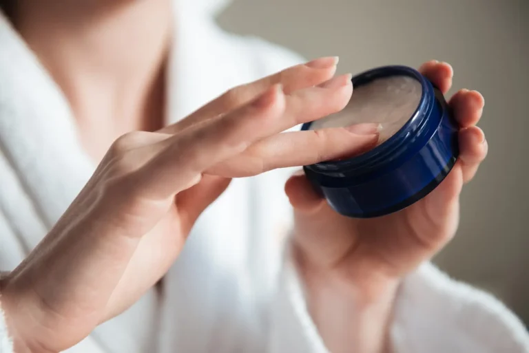 A person in a white robe is holding a blue container with cream, using their right hand to apply some onto their left hand. The focus is on the hands and the container.