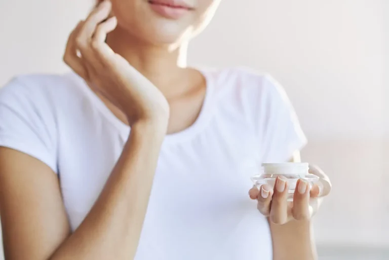 A person in a white t-shirt gently touches their face while holding a small jar of cream. The background is softly blurred, emphasizing a serene and skincare-focused moment.