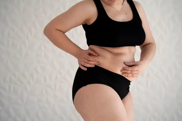 A woman in a black sports bra and underwear stands against a white textured wall, touching and gently holding her stomach with both hands. Her face is partially out of frame.