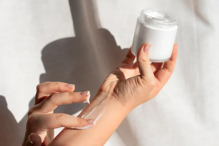 A person is applying white cream from a small jar onto their forearm with two fingers. The background is softly lit, creating a calm and simple atmosphere.