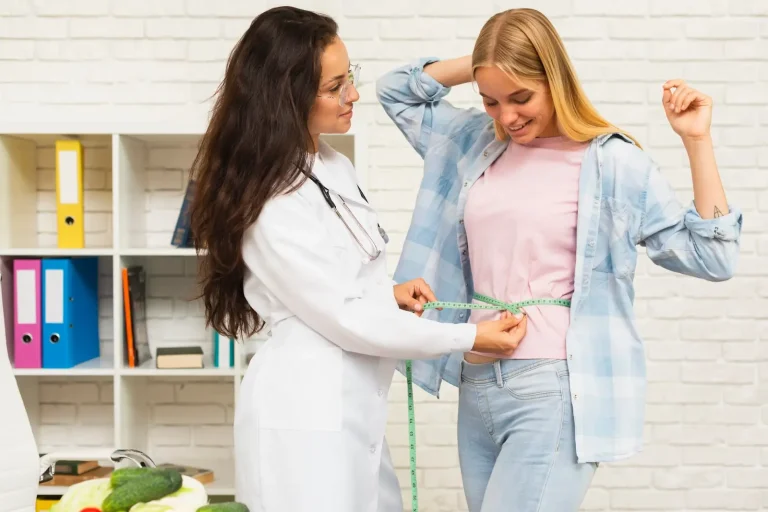 A female doctor measures a young woman's waist with a tape measure. The woman smiles and lifts her arm as the doctor records the measurement. Vegetables and files are on a table in the foreground; shelves and binders are in the background.