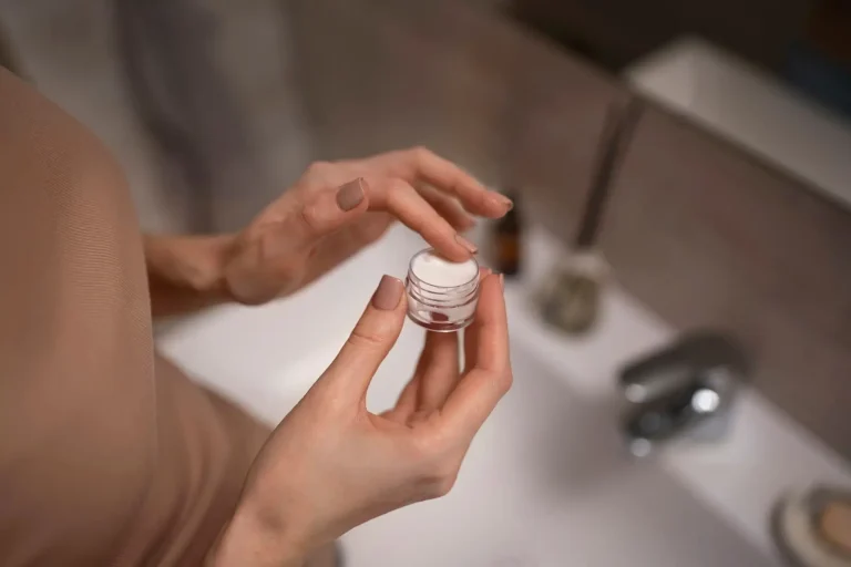A person with painted nails holds an open container of cream in front of a bathroom sink. The focus is on their hands and the small jar, while the blurred background shows a faucet and toiletries.