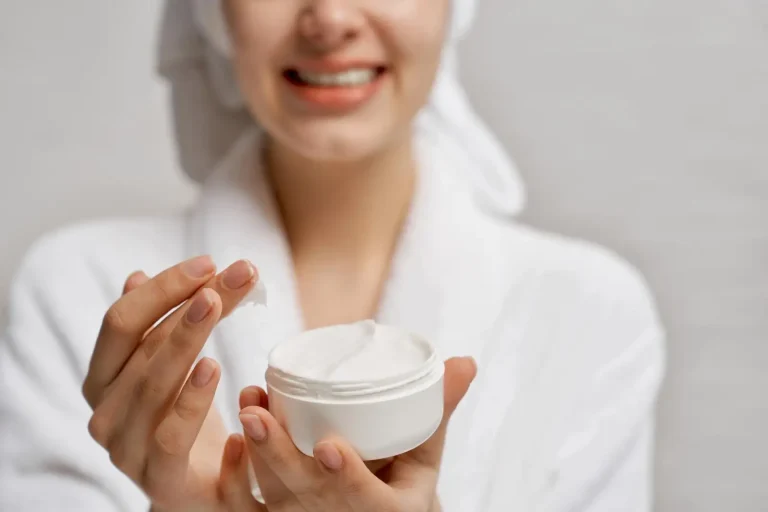 A person in a white bathrobe and a towel wrapped around their head holds a jar of white cream, smiling softly. Their fingers are poised to scoop some cream from the open jar. The background is softly blurred.