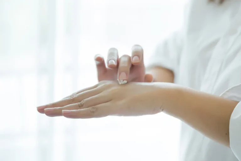 A person applying lotion to the back of their hand with a gentle touch. The background is softly blurred, creating a calm and soothing atmosphere. The sleeves of a light shirt are visible, suggesting a casual setting.