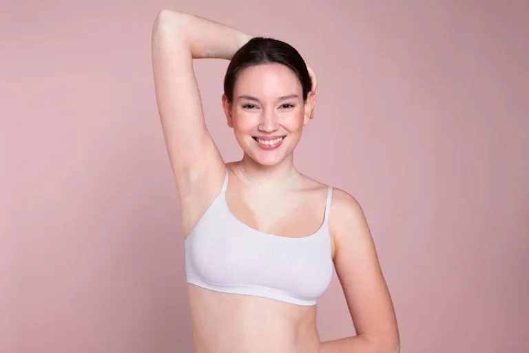 A person with a friendly smile poses against a soft pink background. They are wearing a light gray tank top and have one arm raised behind their head, revealing a natural underarm.