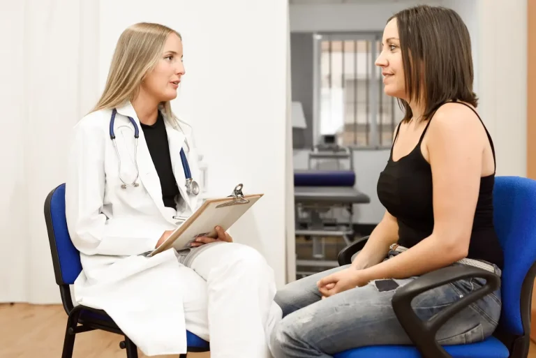 A female doctor in a white coat with a stethoscope talks to a woman in casual clothes, holding a clipboard. They are seated facing each other in a medical office.