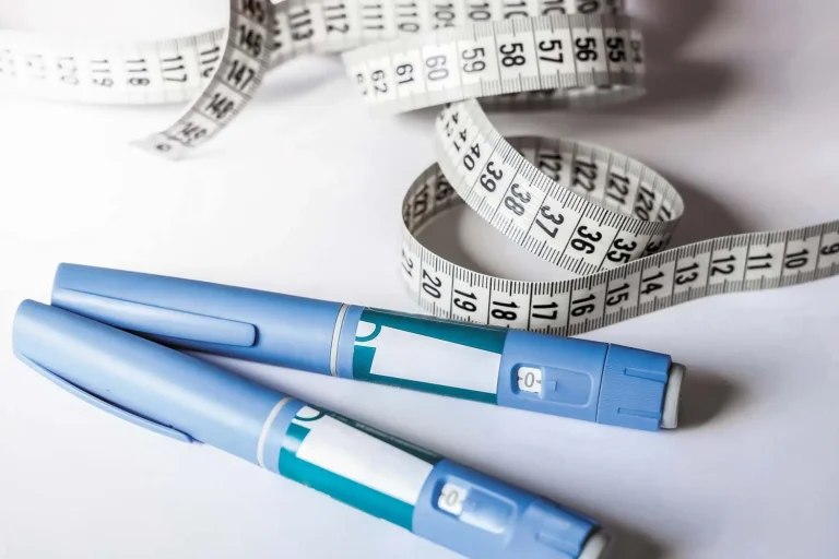 Two blue medical injection pens lie on a white surface next to a coiled white measuring tape with black numbers, suggesting a theme of weight management or diabetes care.