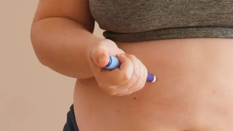 A person wearing a gray top gives themselves an injection in the abdomen using a blue insulin pen. The focus is on the hand holding the pen and the stomach area.