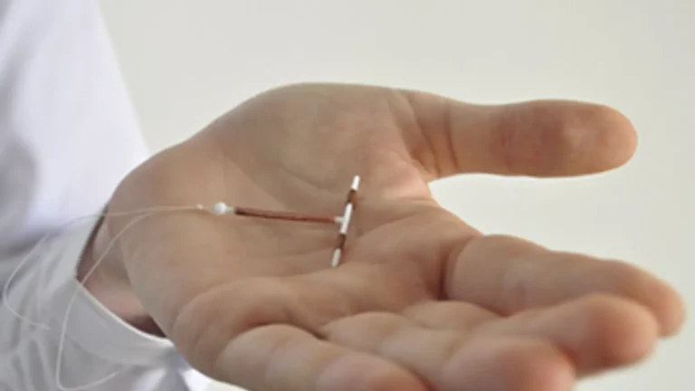A close-up of a person's hand holding an intrauterine device (IUD), a small T-shaped contraceptive with copper wire and white plastic parts, against a plain background.
