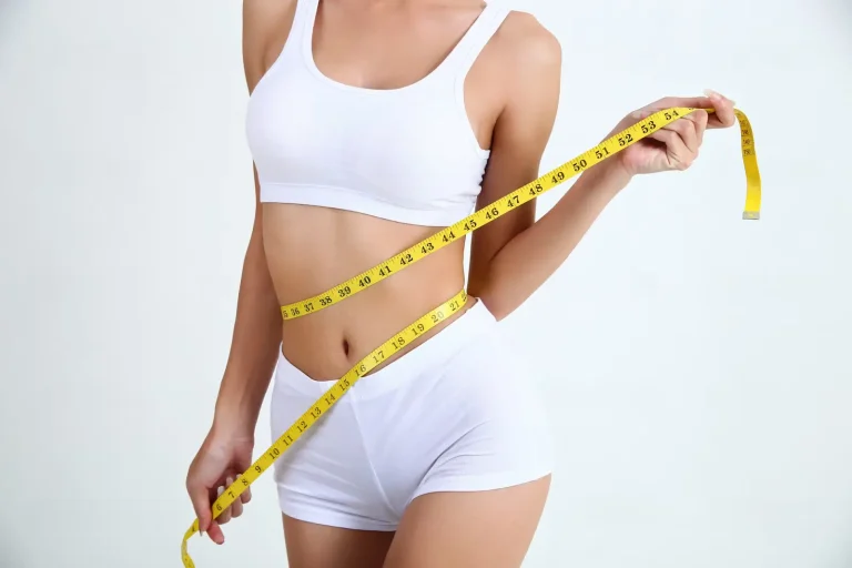 A woman in white underwear holds a yellow measuring tape around her waist, highlighting body measurements or weight loss, against a plain light background.