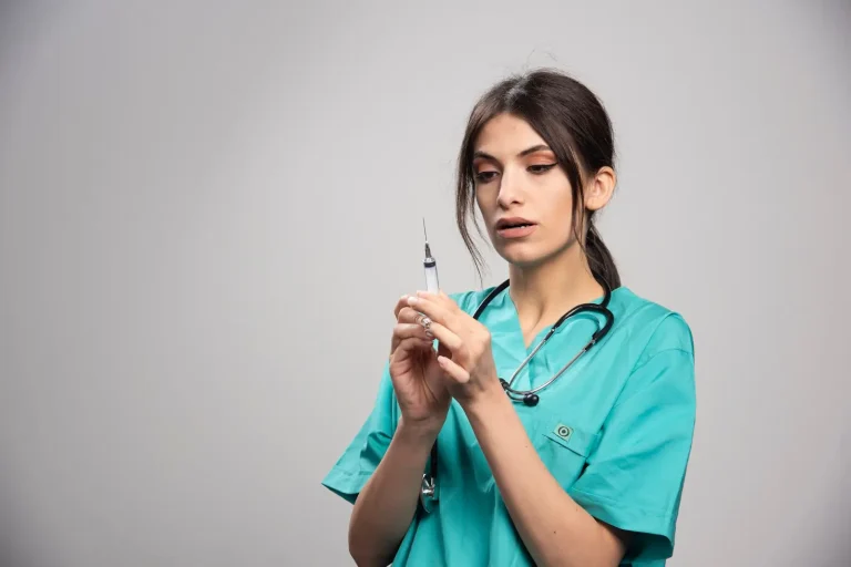 A healthcare professional wearing teal scrubs and a stethoscope prepares a syringe, holding it upright and inspecting the needle against a plain gray background.