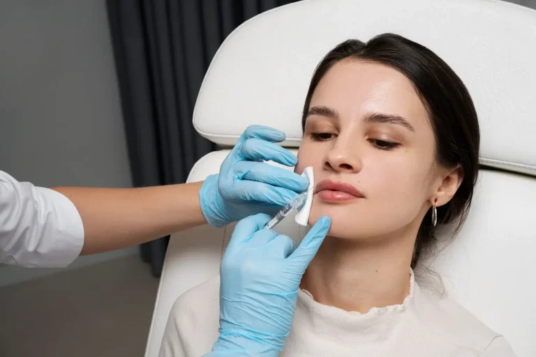 A woman sits in a clinic chair while a medical professional wearing blue gloves injects her upper lip with a syringe, suggesting a cosmetic procedure such as lip filler.