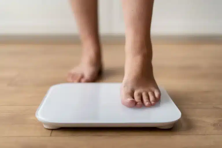 A person placing one bare foot on a white digital bathroom scale on a wooden floor, preparing to weigh themselves.
