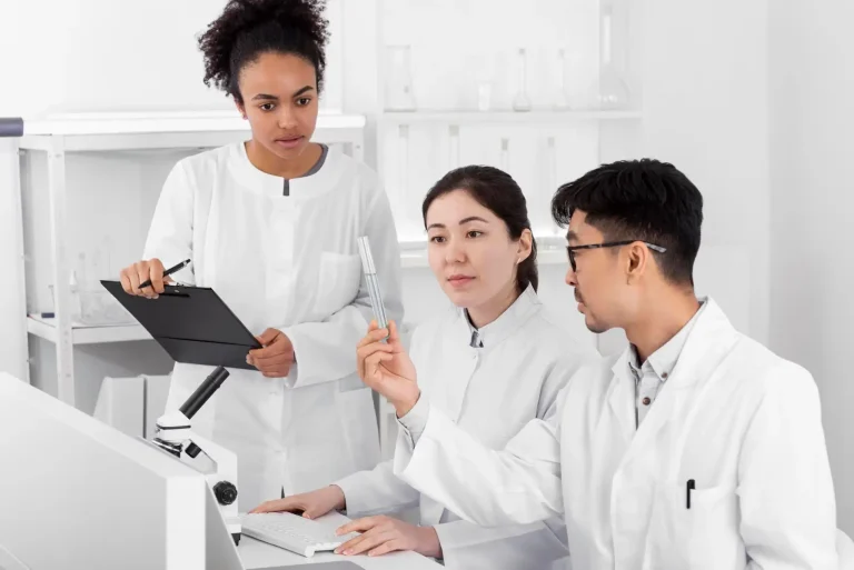 Three scientists in white lab coats work in a laboratory. One holds a clipboard, another types on a keyboard, and the third examines a test tube. A microscope and computer monitor are on the desk in front of them.