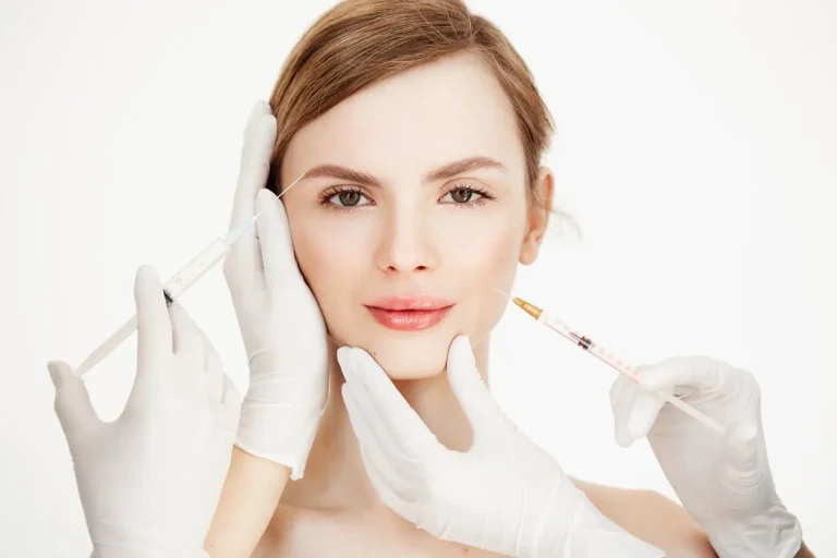 A woman with light skin and brown hair faces forward while gloved hands hold syringes near her face, preparing for cosmetic injections. The background is white and minimalistic.