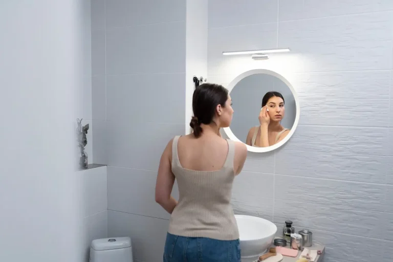 A woman stands in a modern bathroom, looking at herself in a round mirror above a white sink while applying makeup. Various toiletries and beauty products are arranged on the counter.