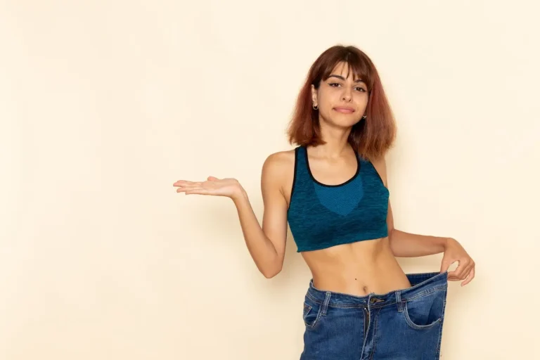 A young woman in a sports bra holds out the waistband of oversized jeans with one hand and gestures with the other, suggesting significant weight loss. She stands against a plain beige background.