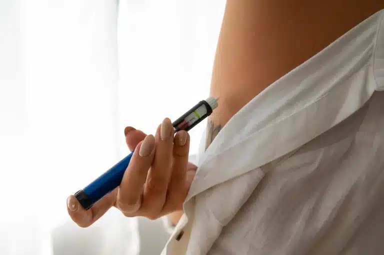 A person wearing a white shirt injects themselves in the abdomen with a blue insulin pen, managing their diabetes. The scene is well-lit with soft, natural light in the background.