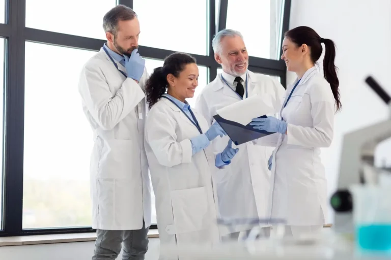 Four scientists in lab coats and gloves stand together in a bright laboratory, smiling and discussing documents or data in a folder, with laboratory equipment visible in the foreground.