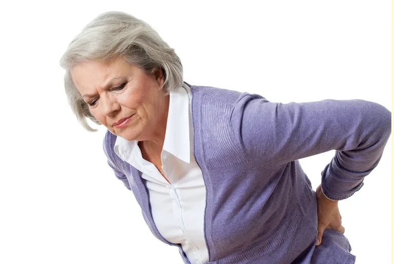 An older woman with gray hair and a purple sweater holds her lower back and appears to be in pain, suggesting discomfort or back pain, against a plain white background.