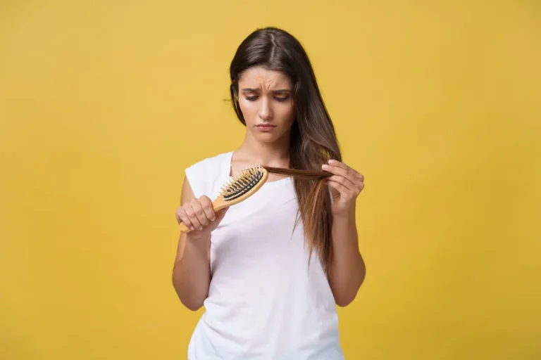 A young woman with long brown hair wearing a white shirt looks concerned while examining hair on a brush and holding a section of her hair, standing against a yellow background.