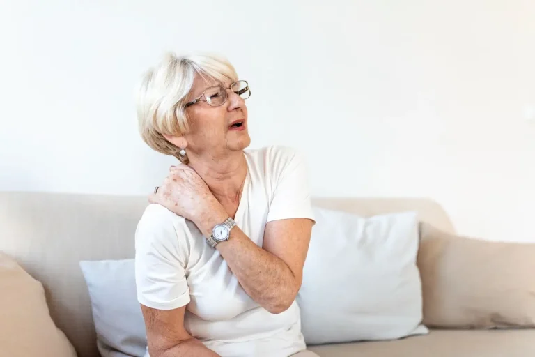 An older woman with short blonde hair and glasses sits on a couch, holding her left shoulder and appearing to be in pain or discomfort. She is wearing a white shirt and a watch.
