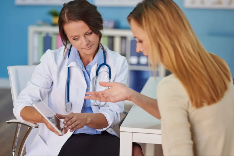 A female doctor with a stethoscope discusses information on a digital tablet with a female patient in an office setting. The patient gestures with her hand while listening attentively.