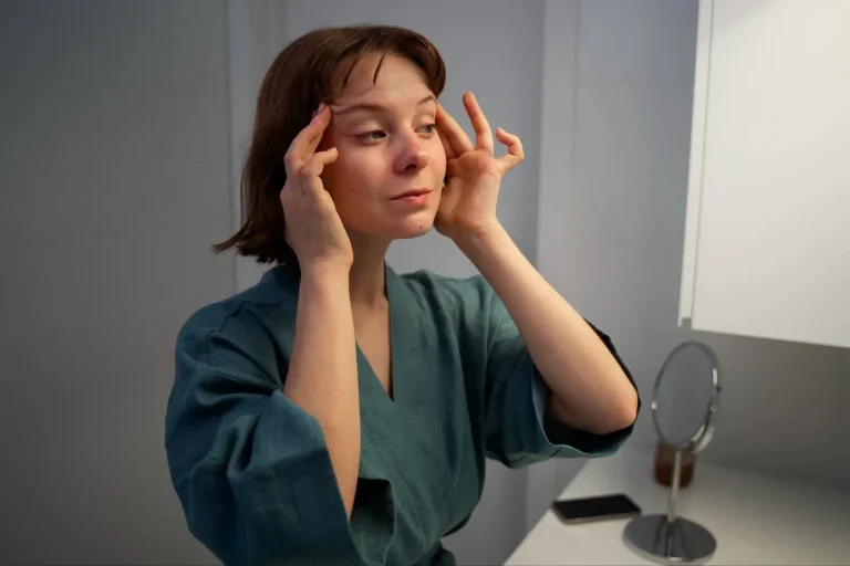 A woman in a teal robe stands in a bathroom, gently touching the skin around her eyes with both hands while looking slightly upward. A small mirror and a phone are on the counter beside her.