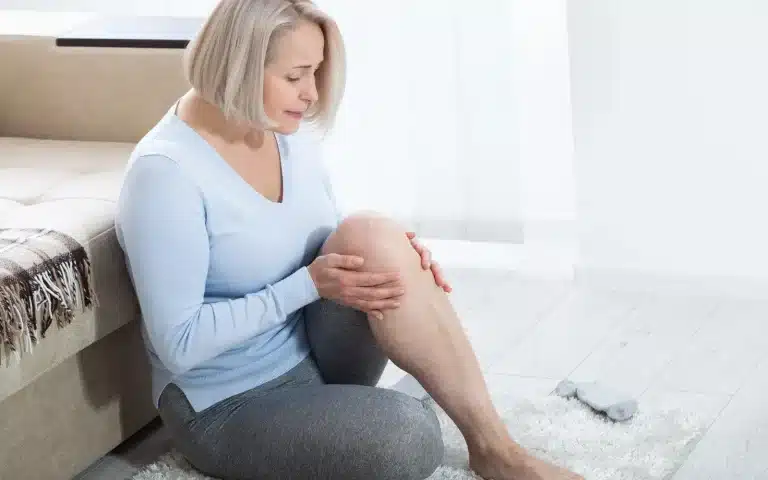 A woman with short blonde hair sits on the floor indoors, holding her knee and calf with both hands, appearing to be in discomfort or pain. She is wearing a light blue sweater and gray pants.