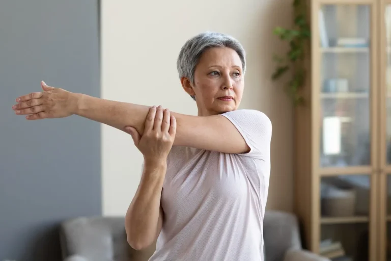 An older woman with short gray hair stretches her right arm across her chest while standing indoors, looking to the side. She is wearing a light-colored short-sleeve shirt.