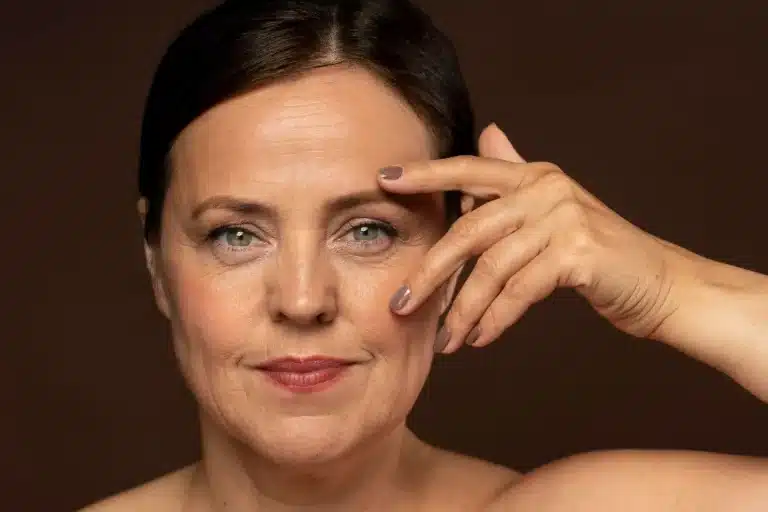 A middle-aged woman with light eyes and brown hair, holds her hand to her forehead. She has a calm expression, subtle makeup, and natural wrinkles, against a plain brown background.