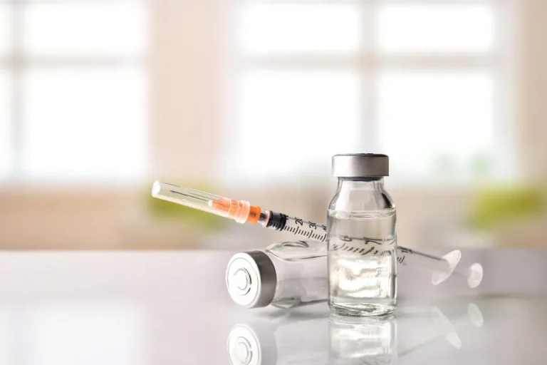 Two glass vials filled with clear liquid and a syringe rest on a reflective surface, with a bright, blurred indoor background.