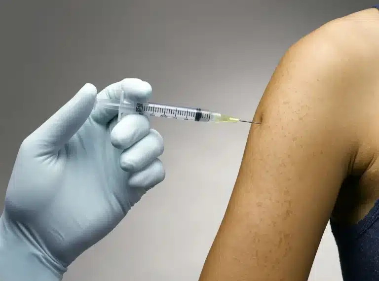 A gloved hand holds a syringe and administers an injection into a person's upper arm. The background is plain and neutral.
