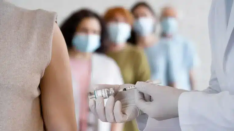 A healthcare worker wearing gloves prepares a syringe with a vaccine as a person waits for an injection. In the background, four people wearing face masks stand in line.