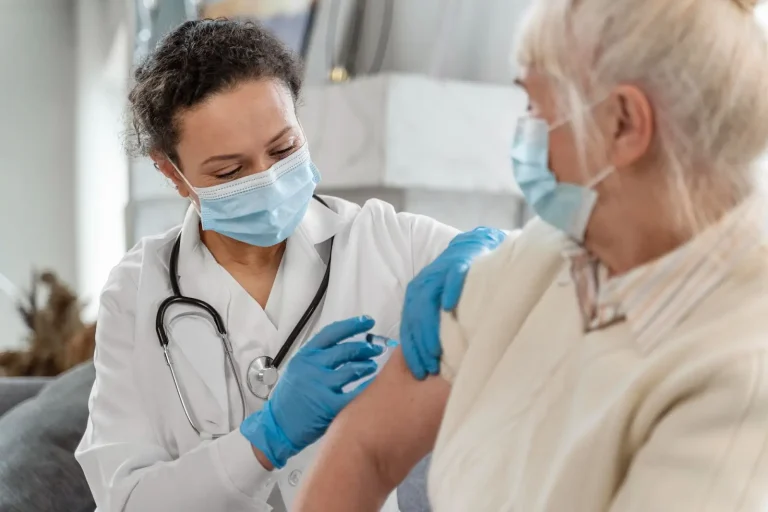 A healthcare professional wearing a mask and gloves prepares to give a vaccine injection to an older woman, who is also masked and has her sleeve rolled up.