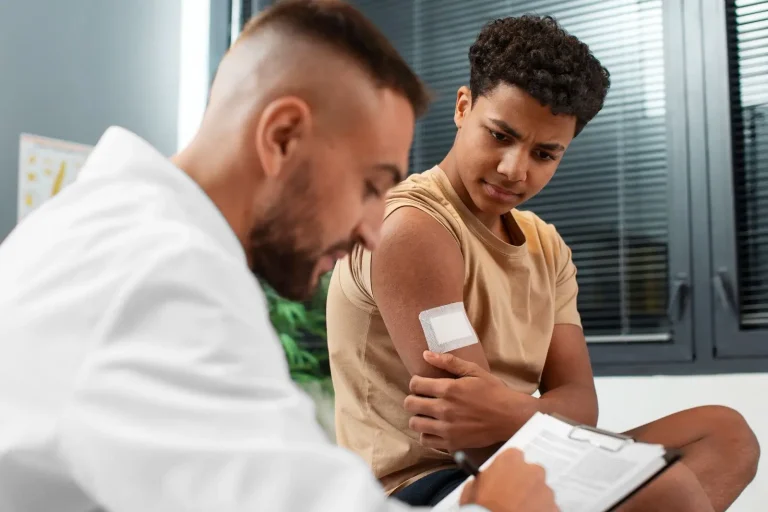 A young person with short curly hair looks at a bandage on their upper arm after receiving a shot, while a healthcare professional in a white coat writes on a clipboard.