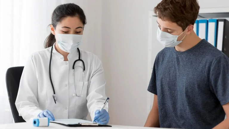 A female doctor wearing a mask and gloves writes on a clipboard while talking to a young male patient, who is also wearing a mask, in a medical office.