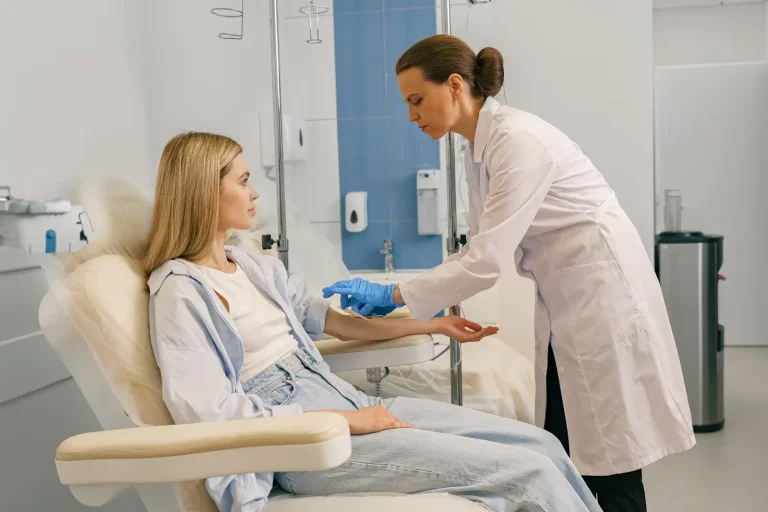 A healthcare professional wearing a white coat and blue gloves prepares to insert an IV into a seated patient’s arm in a medical clinic. The patient sits relaxed in a chair, looking at the nurse.