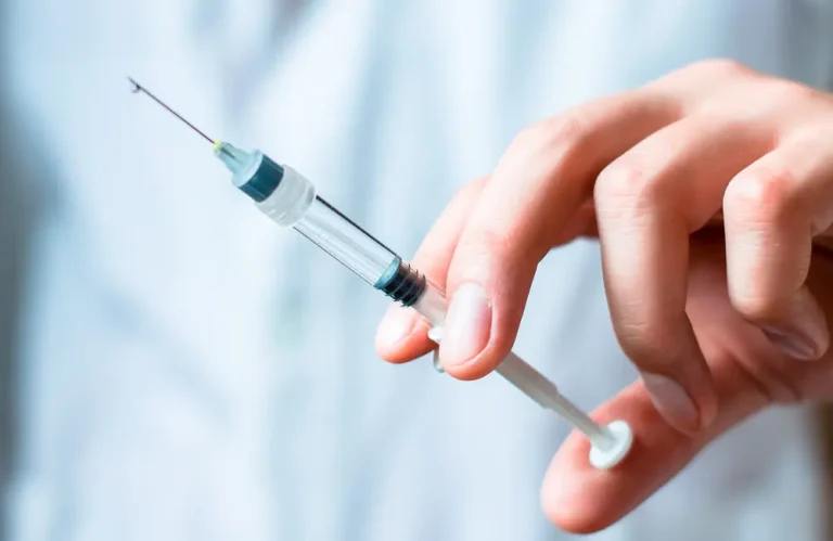 A close-up of a hand holding a filled syringe with the needle pointing upward, ready for an injection. The person is wearing a light blue garment, likely a medical uniform.